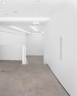 A view through the right window into the gallery, showing a balcony and the top of a staircase headed down to a lower level, in the distance framed artworks in the lower level, and in the foreground on the right on a curved wall is a hanging sculpture.
