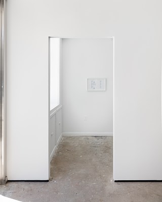 A squared open doorway inside the gallery. In the space beyond the doorway a framed artwork hangs on a white wall.