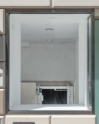 The left window of the gallery’s façade, including the window frame, and a view into the space showing parts of the gate and call buttons of a wheelchair lift.