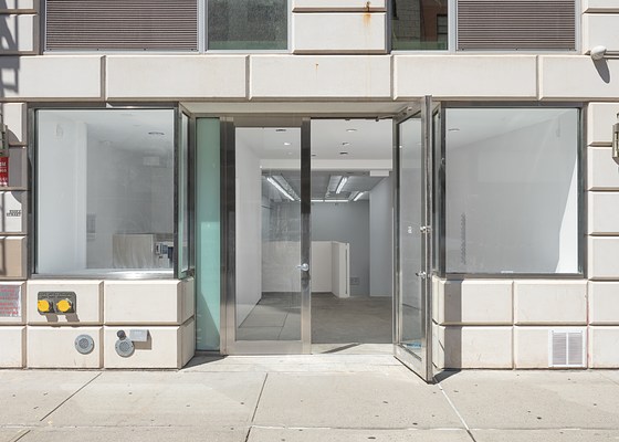 The entire storefront façade of Essex Street Gallery, built of glass and metal trim surrounded by large white bricks, with one of two front doors propped wide open, on a sunny day. On both sides of the open door are the storefront’s large windows showing the interior architecture and some artworks on the walls.