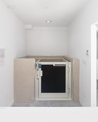 A view through the left window into the gallery, showing part of a framed artwork on the left and the full gate and call buttons of a wheelchair lift in the center and light switches and an alarm code on the right.