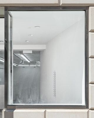The right window of the gallery’s façade, including the window frame, and a view into the space, showing some of the gallery’s overhead lighting fixtures on the left, and on the right on a curved wall is a hanging sculpture.