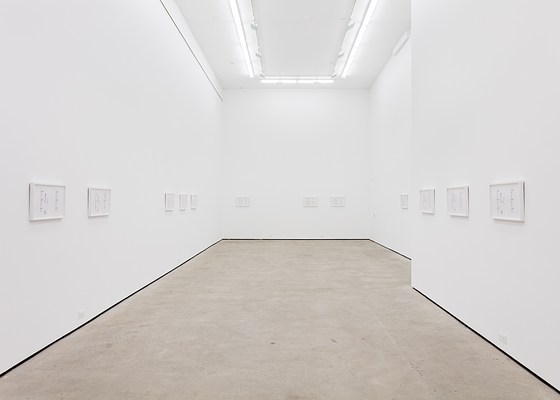 The main space of the gallery with large bright white walls and cool fluorescent lighting. On the walls hang twelve framed artworks unevenly spaced around the walls.