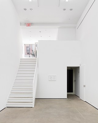 On the left is a large white staircase leading up to the entrance level with windows underneath an EXIT sign and a faint view of a hanging sculpture. In the center is a large white wall with a framed artwork. On the right is a doorway to an elevator lift.