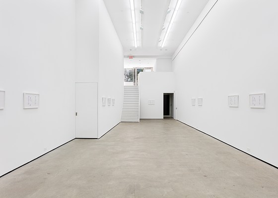 The main space of the gallery with large bright white walls and cool fluorescent lighting. On the furthest wall’s left side is a large white staircase leading up to the entrance level with windows underneath an EXIT sign, on it’s right side is a doorway to an elevator lift. On the walls hang ten framed artworks unevenly spaced around the walls.