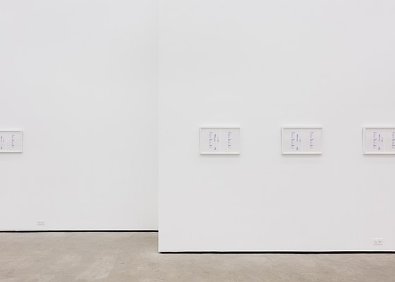A large white wall, interrupted by a break in its center, with four framed artworks unevenly spaced. One of the frames extends outside the picture.