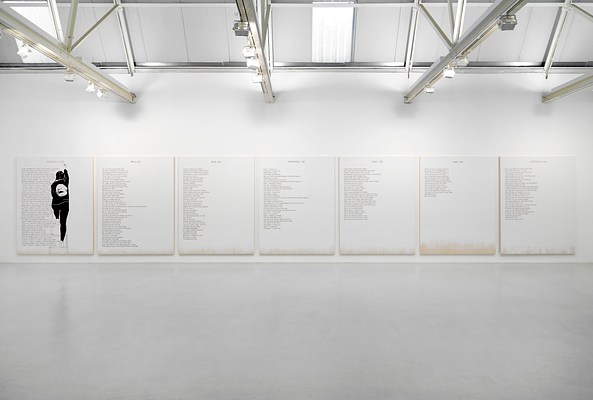 Installation view of a large room with a wide, white wall, light grey ceiling, and light grey, polished floor. We are standing in front of the white wall, which takes up a little over half of the image. Above it, we can see the ceiling which appears to be made out of large, rectangular, and vertically oriented tiles within a structure of steel beams. To the left and in the middle of the ceiling, we can see two rectangular, vertically oriented skylights. Just below the ceiling, perpendicular to the wall, four white beams protrude. On them, we can see spotlights mounted, directed at the wall. On the wall hangs a series of seven paintings, tightly hung in equal intervals. The paintings are white except for around the bottom edges, where we can see the raw cotton canvas. Against the white background, the paintings all have black text in blocks aligned to the left margin, and a title at the top center. On the painting to the very left, we can see the silhouette of a woman with the back against us, painted in black and white. She is standing on a ladder, with her right leg on the first step and her left on the third, while painting the title at the top of the painting with a brush held in her raised right hand.