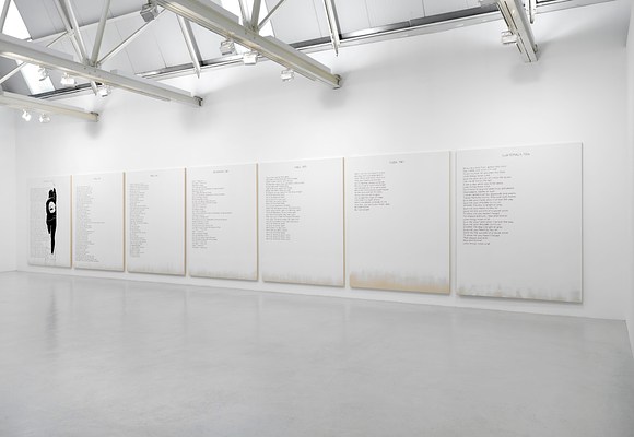 Installation view of the same a large room with a wide, white wall, light grey ceiling, and light grey, polished floor as in the previous image. We are standing to the right in front of the white wall, which takes up a little over half of the image, and at the left edge of the image we can see a corner and a sliver of a perpendicular wall largely out of frame. Above it, we can see the ceiling which appears to be made out of large, rectangular, and vertically oriented tiles within a structure of steel beams. To the left and in the middle of the ceiling, we can see two rectangular, vertically oriented skylights. Just below the ceiling, perpendicular to the wall, four white beams protrude. On them, we can see spotlights mounted, directed at the wall. On the wall hangs a series of seven paintings, tightly hung in equal intervals. The paintings are white except for around the bottom edges, where we can see the raw cotton canvas. Against the white background, the paintings all have black text in blocks aligned to the left margin, and a title at the top center. On the painting to the very left, we can see the silhouette of a woman with the back against us, painted in black and white. She is standing on a ladder, with her right leg on the first step and her left on the third, while painting the title at the top of the painting with a brush held in her raised right hand.