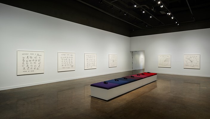 Bench with 7 cushions from purple to blues to reds and 7 headphones surrounded by 6 black and white drawings on 2 walls.