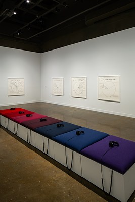 Bench with 7 cushions from purple to blues to reds and headphones sit in the foreground with 4 piechart drawings on 2 walls.