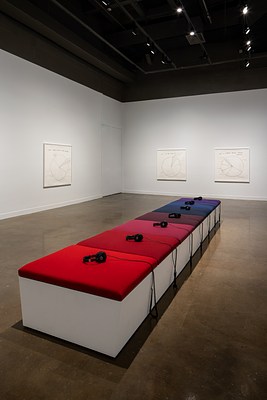 Foreground bench with red, blue and purple cushions and headphones directs viewers to 3 framed pie-chart drawings on 2 walls.