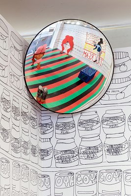 A close up photograph of a round traffic mirror installed in a corner of the gallery below the ceiling. The image shows the corner covered in black and white wallpaper seen throughout the gallery, the mirror and a corner of white ceiling above. In the mirror, the striped laminate floor appears especially bright and distinct. Visible on the floor is the blue plastic milk crate, the leg sculpture and the woman bending down. On the wall reflecting into the mirror we can see the bright red silhouette of legs as well as a large wall work.