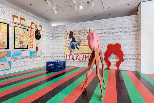 A different installation view of the gallery taken from the left. From this angle we can see that the sculpture of the woman bending down, which we had been directly facing in the last two images, is a cutout made of plywood or another thin material. Only the butt of the woman has any width, being three or four times as thick as the rest of the sculpture. On the far wall, visible from behind the free standing sculpture of the woman bending down, is a red cutout silhouette of the same woman. Only the shape of her body as seen from the front is visible. The cutout shows the top of her rounded butt, the curve of her thighs and knees and calves, and her dainty feet on the floor. To the left of the cutout against the wall is a large painting with the word "ice cream" printed in all caps. On the far left wall is another large wall work. A blue plastic milk crate sits on the striped laminated floor in the corner of the gallery.