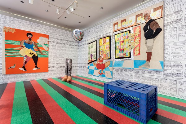 Another installation view of the gallery. From this angle we can see a large orange painting on the back left wall. The painting depicts a person standing against an orange and green background. They have dark skin, an afro, a blue tank top, and colorful patchwork pants. To the right we see a free standing sculpture of legs, a traffic mirror installed above in the corner, two large paintings hung at offset heights and a blue overturned plastic milk crate.