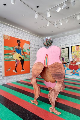 A closer view of the free standing sculpture of a big woman bending down with her head reaching the ground. From this angle we can see that most of the sculpture is a thin cutout made of wood or another thin material. The sculpture is painted with visible brush strokes. The woman's skin is brown, she's wearing a bubblegum pink bathing suit and her vagina, lifted directly toward our view, is painted like a rainbow, starting with red, orange, yellow, blue and green rings. From this view, we can see her legs and vagina and butt while her head is hidden by one calf. Behind the sculpture, the walls are hung with large colorful wall works.