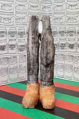 Another detail of the sculpture of legs and feet seen in previous installation images. The legs are loosely formed and are of slightly different size and shape. They appear to be made of lightly painted plaster. The legs are painted thinly with black, much white coming through, and the boots in a light brown. Long, roughly shaped legs end in bulbous brown boots at the floor. At the top of the sculpture the legs end in two rounded mounds, like hip bones between which is painted a white oval which resembles a vagina.