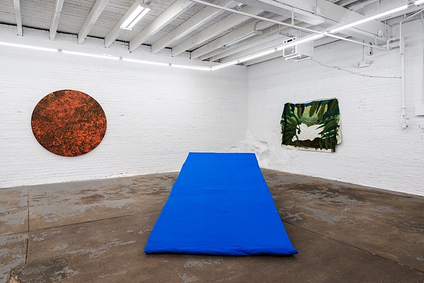 Installation image of Memory of a Body. Left to Right: “Atomic Tangerine: Looking at the Sun with Your Eyes Closed,” “Cerulean: Big Sur Summer 2010,” and “Looking Through the Leaves at Two Lovers Making Out.” This picture was taken at eye level height facing a corner of Mother Gallery in Beacon New York. The floor looks rusty with splotches of burnt coppers, dirty browns and exposed grays. The walls are brick, painted with so many coats of white paint that the definition of each brick is hard to distinguish, making individual bricks unclear or hard to define. Hanging from the ceiling are tubes of fluorescent lightbulbs, emitting a crisp and clear white light.. . In the center of the image is a large blue shape, it looks like a wedge or ramp on the floor. To the left of the blue wedge, hanging on the wall behind is a large circle with splotches of oranges, browns, and gold. To the right of the blue wedge, on the wall catty corner to the orange circle is a rectangular canvas that is mostly green in color with a white shape in the center of the canvas.