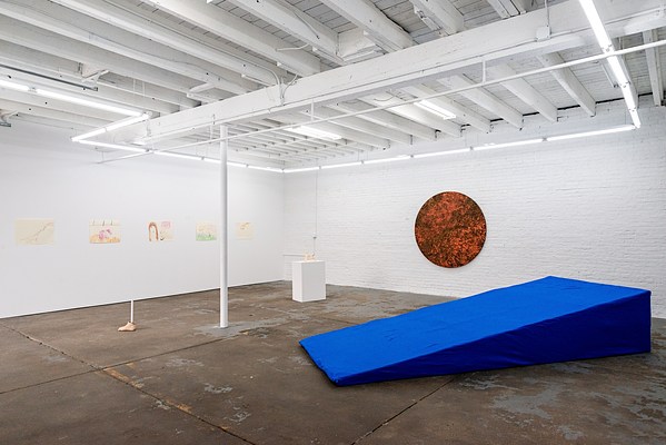 Installation image in which “Atomic Tangerine: Looking at the Sun with Your Eyes Closed” hangs prominently on the wall just behind the blue wedge. A circular painting 64 inches in diameter, it is slightly smaller than the average human arm span. Instead of using canvas, Emilie applied oil and enamel paint to a gold cotton cloth. The way these materials interact creates a dynamic surface and the gold cotton underneath creates additional flickers and sparkles of light that reflect as your eye moves around the painting. The surface also has a visual quality like fire or embers because there is a mix of warm colors like deep reds, dirty oranges, and burnt sienna in a variety of shades, some are bright and some are dark. More literally, the surface looks like a galaxy of different shades of orange, browns, and golds that are speckled, splotched, and colliding with one another across the composition.