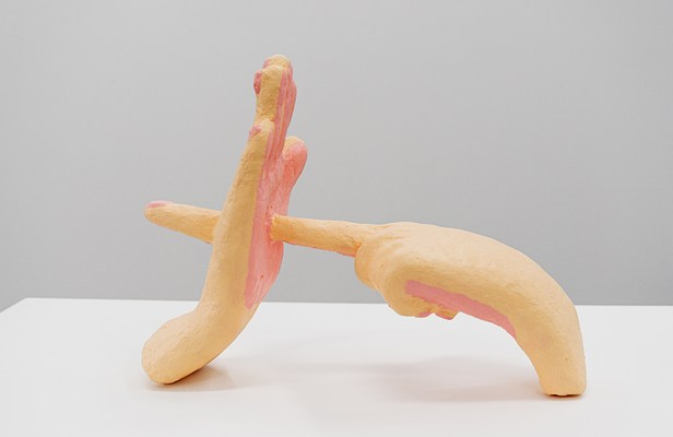 “Finger Through Palm” is a small papier-mache sculpture of two hands cut off at the wrist. It is painted thickly with cartoony peachy pink skintone, and the fingernails are glossy light pink. The right hand has its palm facing out, with its fingers closed together. The left hand has its index finger extended, poking the center of the palm, and the finger pierces through the palm to the other side.