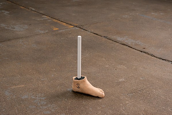 On the floor, is a small, ceramic sculpture of a left foot with a white steel rod sticking out of it, called “Outerspace: My Dad’s Old Tattoo, Smiley Ankle”. The sculpture has been painted to match the color of the artist’s dad’s skintone, which is a light tan. On the ankle, the artist carved out the drawing of a weird smiley face that is an actual tattoo on her father’s foot. The inside is filled with soft, black foam, which seeps out of the incision of the tattoo.