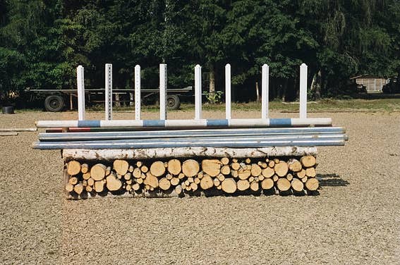 View of a sculpture on a field with trees in the background. The sculpture consists in a rectangular horizontal stack of chopped wood with the round chopped surfaces facing us. Across, on top of the wood lies a thin stem of a birch tree and chromed metal poles. Stacked on these, we can see what appears to be beams and poles from horse jumping obstacles in white with blue stripes. The sculpture sits on a field of what looks like dry grass in a light brown hue. It is sunny and the stack casts short shadows to the right. Behind the sculpture, we can see dark green conifer trees at the edge of the field. In front of the trees to the left, we can see a low trailer with wheels and in the far right we can glimpse a low building. In front of the trailer, behind the sculpture to the left on the grass, we can see two wooden beams lying horizontally in the picture frame.