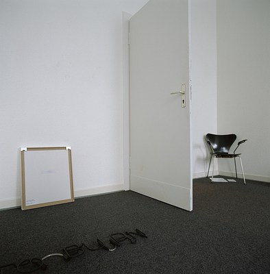 Installation view of room with two white walls and dark grey carpeted floor. The walls form a corner to the upper right in the image. Perpendicular to the left wall, a white door is swung open. It has a metal door handle and juts out into the room. To the left of the door, a square picture stands on the ground, leaning against the wall. It has its back turned against the room and is framed. We can see brown tape along the edges and two metal hangers in each top corner. The back surface is white, and has text in the center and bottom left corner but it is too small for us to discern. In front of it, parallel to the wall on the carpeted floor lies a neon sign consisting in letters. We can see two words, of which the first is mainly out of frame aside from the letter "O." Following this, we can read "burn" in capital letters. The neon letters have been painted in black opaque varnish, and the sign appears to not be lit. In the corner, to the right of the door, stands a black chair with black seating, backrest and handles. It has chrome legs, and stands facing the room. The chair appears to be by a modernist designer such as Jacobsen or a knockoff. Its backrest is triangularly shaped. Underneath the chair on the carpet, we can see a folder to the left and a smaller booklet to the right. In the backrest, light is reflected from a window out of frame.