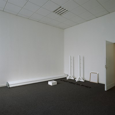 Installation view of the same room with two white walls and dark grey carpeted floor as in the previous image but from another angle. The walls form a corner to the left in the image. Perpendicular to the right wall, at the right edge of the image, a white door is swung open. It has a metal door handle and juts out into the room. To the left of the door, a square picture stands on the ground, leaning against the wall. It has its back turned against the room and is framed. We can see brown tape along the edges and two metal hangers in each top corner. The back surface is white, and has text in the center and bottom left corner but it is too small for us to discern. Next to it, parallel to the wall, stand two white poles from an horse jumping obstacle. Perpendicular to them, alongside the wall to the left, lies a stack of bars belonging to the same obstacle. In front of the two poles we can see a neon sign lying on the floor made up from letters, parallel to the right wall. From out viewpoint, it is not possible to discern the writing but we can see that the letters have been painted in black opaque varnish, and that the sign is not lit. On the floor in front of the sign, we can see a low white cube with a hole in the top at its center. Possibly, this is also related to equestrian sports. The ceiling is made up of white square tiling, and directly above the cubicle, an embedded, gridded light fixture in silver is visible. . . . 