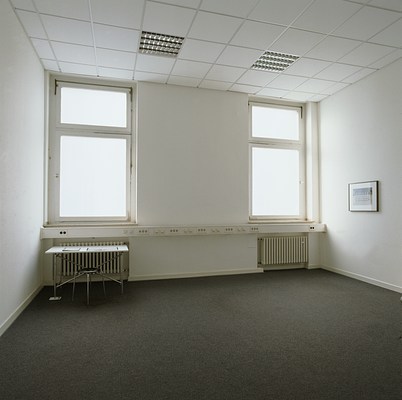Installation view of a white room with three walls, white tiled ceiling, and dark grey carpeted floor. At the edges of the wall facing us, we can see two identical windows. The windows are rectangular and reach all the way to the ceiling. They are divided into two window panes, the lower twice as large as the upper. Directly beneath the windows, across the entire wall, runs a white bar with light switches and outlets. Under both windows, below the bar, we can see white rectangular radiators. Through the windows we can see white light. The ceiling is made up of white square tiling, and two embedded, gridded light fixtures in silver are visible. On the floor in front of the left window stand a rectangular desk and a round stool. The table frame is made from chromed metal. The table top is a white board. The stool has a black seat and its chromed legs match the table frame. On the table lies two folders with black frames next to each other. The left one is closed and the right one is open. On the wall to the right of the windows we can see a rectangular photograph in landscape orientation, framed by a white passepartout frame and black thin frame. The photo appears to be of a sculpture constructed from chopped wood onto which metal elements and horse jumps have been stacked. The photo and the wall around it is illuminated by light white emanating from the nearby window.