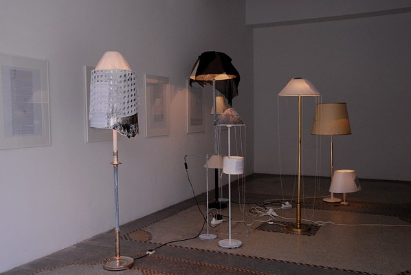 A darker installation view of a gallery. Along the gallery wall four framed texts hang. Floor lamps and desk lamps sit on the gallery floor in a crowd of different styles and heights. Many of their shades are draped with scarves and some of the lamp shades have texts attached. The lamps are on but they're not throwing much light into the space.