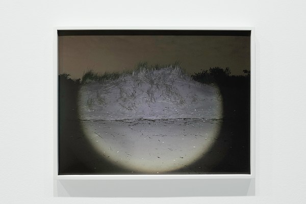 A semi-circle of light illuminates a sand dune against a sepia-colored sky in a framed photograph on a white gallery wall.