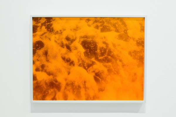 Framed orange-colored photograph of foam from ocean waves hangs on a white gallery wall.