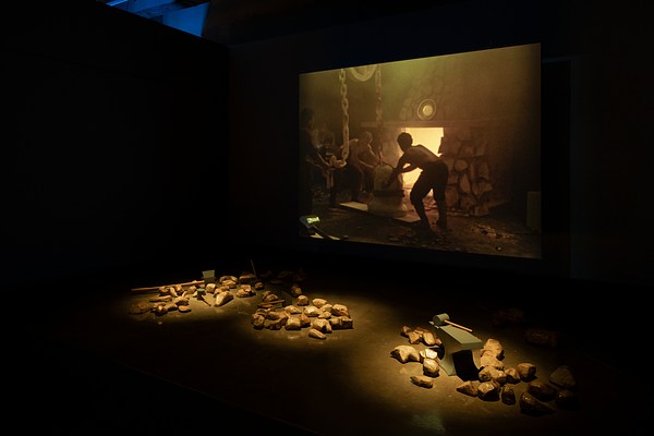 Three piles of gold-painted rocks and mallets are grouped in front of a screen on which an image of a man working in a forge.