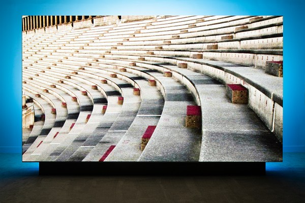 A large screen with the projected image of convex concrete stadium seating stands in the Gallery, backlit by a blue light.