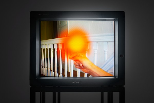  A hand and arm  extended into an interior  stairway with white railing and  balustrades. A bright, flamelike, over-exposed object sits  atop the hand.
