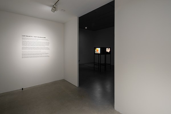 Two rooms, one illuminated and one  in darkness, with text on the wall of the first  room and videos on monitors in the other.
