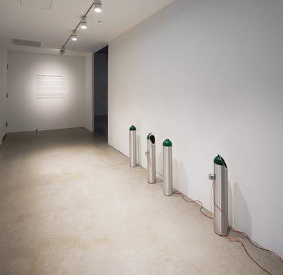On the right side of a hallway sit 4  cylindrical tanks with painted green tops, some  with holes and wires. Text on back wall.