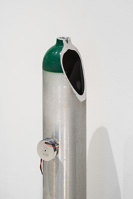 Thin silver oxygen tank with a  painted green top. A portion of the right  side of the tank is removed. A  transducer with wires is attached to the  left side.