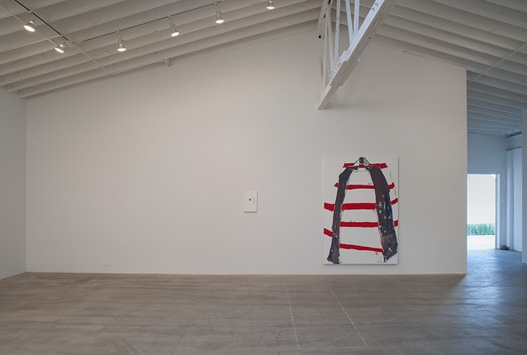 Installation view of a gallery space with a white wall and grey, concrete floor. Above the wall, we can see a slanted ceiling. In the ceiling, white beams run parallel to the white wall, and where they meet slightly off center to the right in the image, a large white beam protrudes into the room perpendicular to the wall. To the left in the ceiling, we can see five spot lights. On the wall, we can see two paintings hanging below the large beam. To the left, we can see a small-scale white painting in portrait format with a piece of cloth attached to the surface. To the right, we can see a large-scale abstract painting with red and grey shapes against a white background. To the right of the wall, the room continues further back into the image, where we can see a doorway. Through the doorway, we can see daylight coming in, and glimpse a courtyard with green grass and a light grey wall. The concrete floor reflects some of the light.