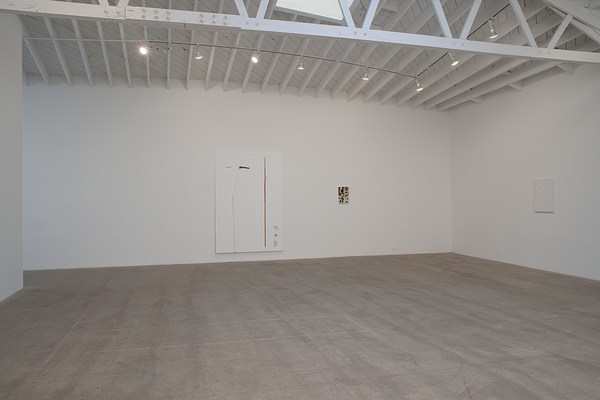 Installation view of a gallery space with two white walls, and concrete grey floor. The left wall takes up most of the upper half of the image, and meets the right wall in a corner to the far right in the image. In the center of the left wall, we can see a large, white rectangular painting in portrait format with think wooden sticks attached to the surface. To the right of the large painting, we can see a smaller painting in portrait format in colorful abstract pattern. On the right wall, we can see a medium sized white painting in portrait format with a backlit photo slide mounted to the surface. In the ceiling, we can see a white, large horizontal beam with beams in a zig zag pattern on top of it. Below this beam, white, narrower beams run vertically, perpendicular to the left wall. We can also see a railing with spot lights.