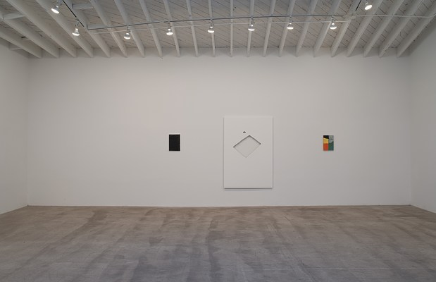 Installation view of a gallery space with a white wall and grey, concrete floor. On the white wall, we can see three paintings hung with equal space between them. They are all rectangular, and in portrait format. The left one is a small-scale black monochrome, the middle one a large white painting with a cut-out in the center, and the right one in a small-scale painting with a colorful geometrical pattern. In the ceiling, we can see parts of white beams in vertical and horizontal directions. In the ceiling, we can see white beams running vertically, perpendicular to the  wall. Across these, we can see a railing with spot lights.