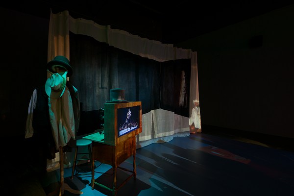 Stage set up featuring a beige curtain, a man’s outfit hung on a coat rack and a wooden desk with a poster on the front and a top hat on the top.&nbsp;
