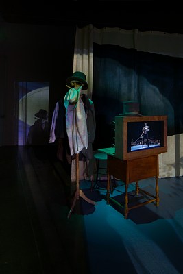 Stage set up featuring a beige curtain, a man’s outfit hung on a coat rack and a wooden desk with a poster on the front and a top hat on the top.&nbsp;