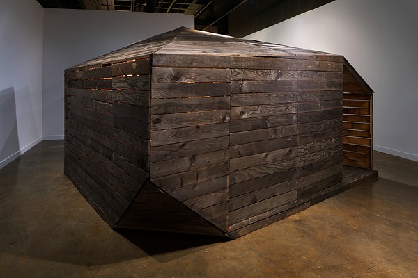 A large dark slatted wood cabin structure with pointed roof and lit from within, almost filling the space it’s in.&nbsp;