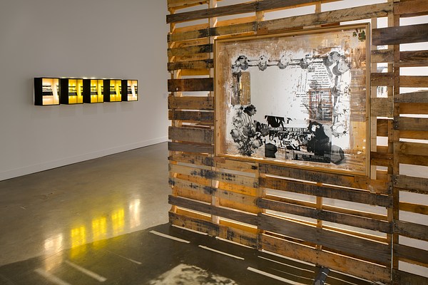 A wood slatted wall with a framed picture on the right, and five dark framed boxes in a row, lit from within, on the far left wall.&nbsp;