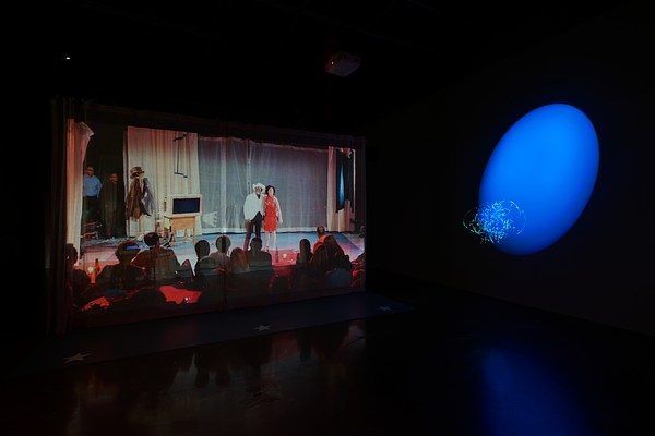 Projection of a stage performance with a man and woman with their arms around each other and a blue spotlight on the right wall lighting up the gallery.&nbsp;