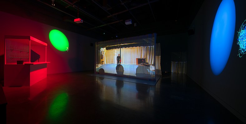 Projection of a man in front of an audience on a large curtain hung in the gallery and a green and blue spotlight projected onto opposite walls lighting the gallery.&nbsp;