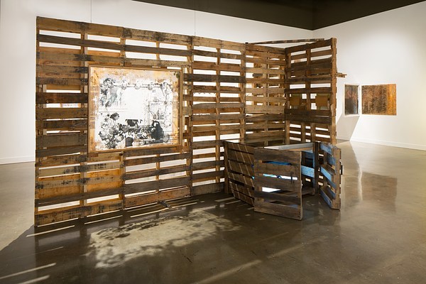 Installation view with a large sculpture in the center of the room made of wood pallets with a large sketch hung on the front facing side and a gate-like arrangement of the wood on the right.&nbsp;
