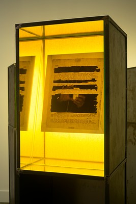 Detailed view of a lit light box with a sheet of paper in each that has certain parts of the text blacked out and an image on the background of the paper of someone taking a photo with a small cell phone.&nbsp;