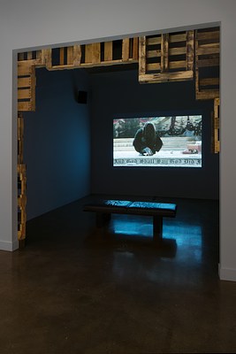 Entry to a gallery room with wood pallets hanging on the top and edges of the wall entrance with a large projector screen on the back wall of the room and a small black bench in front of it.&nbsp;