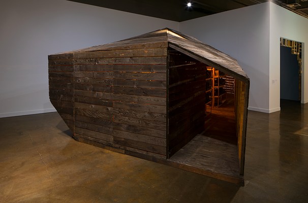 A large dark slatted wood cabin structure with a pointed roof and an open door on the right that leads into the space with warm light coming from within.&nbsp;